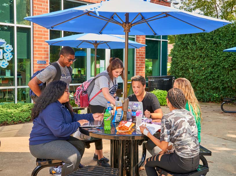A group of students work on separate devices as they sit at a table under an umbrella outside of the Student Community Center on Penn State Greater Allegheny's campus.