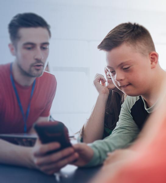 A man and young boy sitting at a table both looking at a cell phone, being held by the man on the left of the photo.